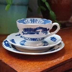 Op de afbeelding: Een witte theekop en schotel set met blauwe draken- en bloemmotieven. De theekop heeft een drakenontwerp, terwijl de schotel bijpassende patronen heeft. De set staat op een houten oppervlak, wat een vintage of antieke uitstraling geeft.