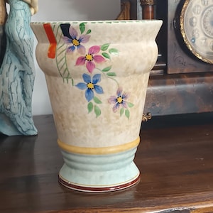 Pode incluir: Um vaso de cerâmica vintage com acabamento bege salpicado, adornado com flores pintadas à mão em rosa, azul e roxo. O vaso tem uma base verde claro e uma faixa amarela. Uma faixa vermelha e preta acentua o topo. O vaso tem cerca de 18 cm de altura.