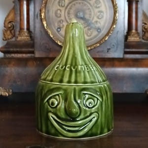 May include: A green ceramic cucumber-shaped jar with a smiling face design. The word "CUCUMBER" is printed on the jar. The jar is sitting on a wooden surface.