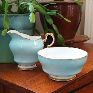 Puede incluir: Un juego de té azul claro con una jarra de crema y un azucarero. El juego tiene un borde dorado y está sobre una superficie de madera.