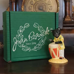 May include: A green box with the text "John Beswick Since 1894" and a figurine of a black cat playing a drum. The cat wears a yellow shirt and white trousers. The drum is orange and red.