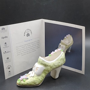Op de afbeelding: Een groene keramische schoen met een witte strik en bloemmotieven. De schoen is versierd met vergeet-me-nietjes en heeft een hoge hak. De schoen maakt deel uit van een collectie van Royal Stratford English fine bone china.