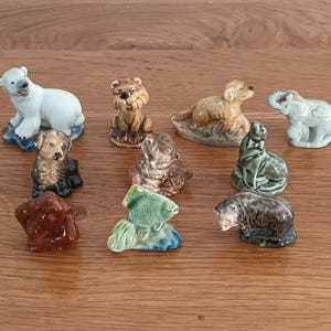 May include: A collection of ceramic animal figurines, including a polar bear, bear, dog, cat, elephant, seal, fish, and beaver. The figurines are painted in various colors and sit on a wooden surface.