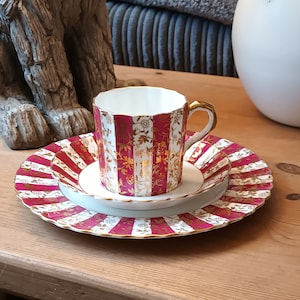 May include: A white teacup and saucer set with a pink and gold floral pattern. The cup has a scalloped rim and a handle. The saucer has a scalloped edge and a pink and gold stripe pattern.