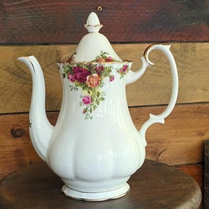 Puede incluir: Tetera de porcelana blanca con ribete dorado y un patrón floral con rosas. La tetera tiene una tapa con un pomo en la parte superior y un asa curva.