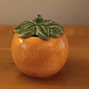 Peut inclure: Un pot en céramique en forme d'orange avec un couvercle en forme de feuille verte. Le pot est de couleur orange vif avec une surface texturée. Le couvercle est vert brillant, ajoutant un élément de contraste. Un objet de décoration unique.