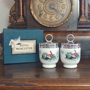 May include: Two white porcelain egg coddlers with chrome lids and ring handles, featuring a colourful bird and foliage illustration. The set includes a blue cardboard box with a label reading Royal Worcester in black text on a white background.