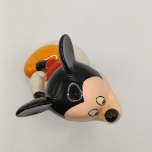 May include: A black, white and orange ceramic figurine of Mickey Mouse. The figurine is lying on its side with its head tilted to the side. The figurine has black ears, a black nose and black eyes.