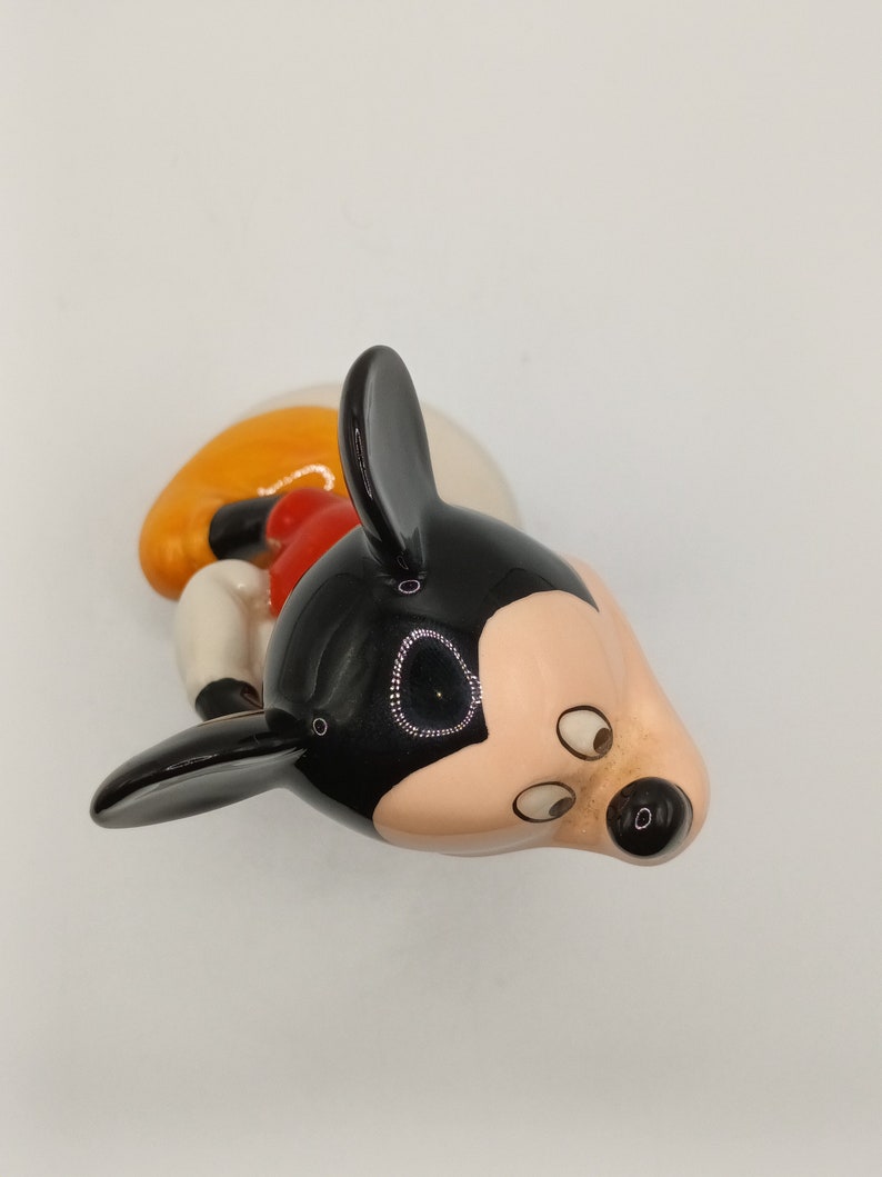 May include: A black, white and orange ceramic figurine of Mickey Mouse. The figurine is lying on its side with its head tilted to the side. The figurine has black ears, a black nose and black eyes.