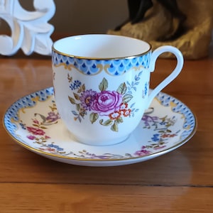May include: A white porcelain teacup and saucer set with a floral pattern in shades of pink, purple, and blue. The cup and saucer have a gold trim and a blue and gold scalloped design around the rim.