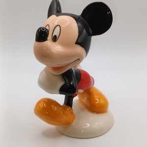 May include: A ceramic figurine of Mickey Mouse. He is standing with his right foot forward and his left arm bent at the elbow. He is wearing his signature red shorts with a yellow button and black shoes. His ears are black and his face is a light peach colour.
