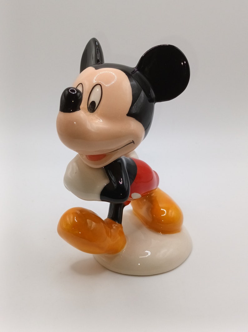 May include: A ceramic figurine of Mickey Mouse. He is standing with his right foot forward and his left arm bent at the elbow. He is wearing his signature red shorts with a yellow button and black shoes. His ears are black and his face is a light peach colour.