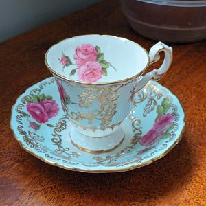Puede incluir: Un delicado juego de taza y platillo de té con fondo azul claro y ribete dorado. El juego presenta un diseño floral con rosas rosas y hojas verdes.