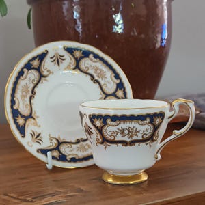 Puede incluir: Un juego de taza y platillo blancos con ribetes dorados y diseños florales azules y dorados ornamentados. La taza tiene un asa pequeña y una base dorada. El platillo es redondo y tiene un diseño a juego. El juego se exhibe sobre una superficie de madera.