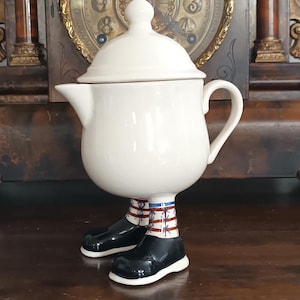 Puede incluir: Una tetera caprichosa de cerámica blanquecina con tapa, boquilla y asa. La tetera está sostenida por zapatos y piernas negros con calcetines a rayas rojas, blancas y azules. Un artículo decorativo único.