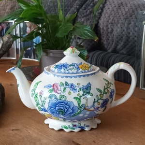 Op de afbeelding: Witte keramische theepot met een bloemmotief in blauw, roze en geel. De theepot heeft een gebogen tuit, een handvat en een deksel met een decoratieve knop. Het ontwerp bevat een blauw bloemmotief langs de rand.