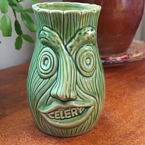 Può includere: Un vaso in ceramica verde chiaro a forma di gambo di sedano con una superficie strutturata a linee verticali. Il vaso presenta un volto scolpito con sopracciglia prominenti, occhi circolari e una bocca larga con la parola CELERY in rilievo.