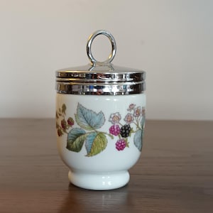 Puede incluir: Un huevero de cerámica blanca con tapa y asa plateadas. El huevero está decorado con un patrón floral de moras y hojas.