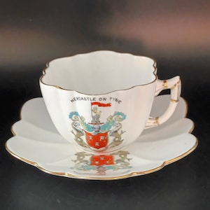 May include: A white teacup and saucer with a gold rim. The cup has a crest with the words "Newcastle on Tyne" and a red flag.