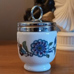 Peut inclure: Un coquetier en porcelaine blanche avec un motif floral bleu et vert et un couvercle argenté avec une anse en forme d'anneau.