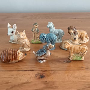 May include: A selection of miniature ceramic animal ornaments. The collection includes a deer, owl, hedgehog, duck, elephant, horse, lion, farmer, and cow. The figurines are painted in various colours and are displayed on a wooden surface.