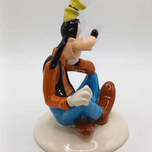 May include: A ceramic figurine of Goofy sitting with his legs crossed, wearing a yellow hat, orange shirt, blue trousers, and brown shoes. He has a black nose and white gloves.