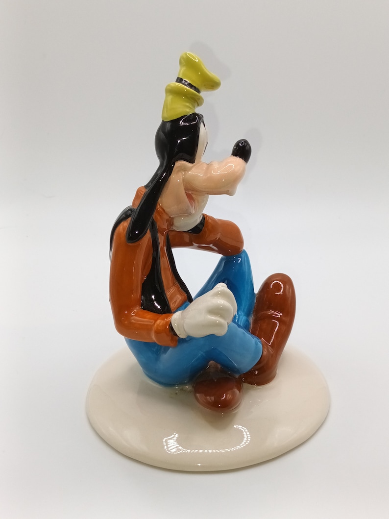 May include: A ceramic figurine of Goofy sitting with his legs crossed, wearing a yellow hat, orange shirt, blue trousers, and brown shoes. He has a black nose and white gloves.