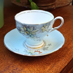 Puede incluir: Un delicado juego de taza y platillo de té con una base azul claro y un patrón floral en tonos grises, amarillos y verdes. La taza tiene un borde dorado y el platillo tiene un borde dorado.