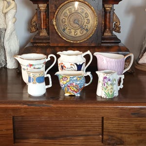 Könnte beinhalten: Eine Sammlung von sechs kleinen, dekorativen Keramikkännchen mit verschiedenen floralen und gemusterten Designs. Die Kännchen sind in Weiß-, Blau-, Rosa- und Gelbtönen gehalten. Sie stehen auf einer dunklen Holzoberfläche, mit einer Vintage-Uhr im Hintergrund.