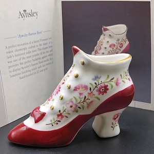 Op de afbeelding: Een witte porseleinen schoen met rode afwerking en bloemmotieven. De schoen is versierd met gouden accenten en heeft een knoopdetail aan de zijkant. De schoen is een decoratief stuk en maakt deel uit van een collectie.