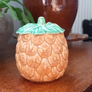 May include: A ceramic pineapple-shaped jar with a lid. The jar is a warm orange colour with a textured surface, and the lid is green, resembling leaves. The jar is on a wooden surface.