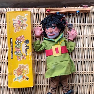 Puede incluir: Una marioneta Pelhams vintage con una túnica verde, cinturón rojo y zapatos negros, acostada sobre una cesta de mimbre. La marioneta tiene una cara pintada y cabello negro. Una caja amarilla con ilustraciones y el texto "Pelhams Marlborough Wilts".