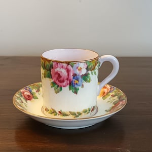 Puede incluir: Una taza de té y un platillo blancos con un patrón floral. La taza y el platillo tienen un borde dorado y presentan flores rosas, rojas, azules y verdes.
