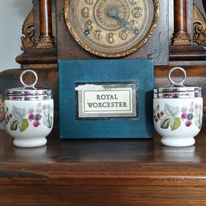 Può includere: Due cuociuova in porcellana bianca con coperchi in metallo argentato e manici ad anello, caratterizzati da un motivo botanico di more. Sono esposti accanto a una scatola color verde acqua con un'etichetta che riporta la scritta Royal Worcester.