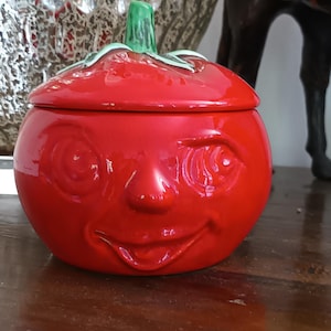 Könnte beinhalten: Ein leuchtend rotes Keramikgefäß mit Deckel in Form einer Tomate mit lächelndem Gesicht. Der Deckel hat einen grünen Stiel als Griff. Die Oberfläche ist glänzend und glatt und zeigt eine runde Gemüsefigur mit geprägten Augen, Nase und Mund.
