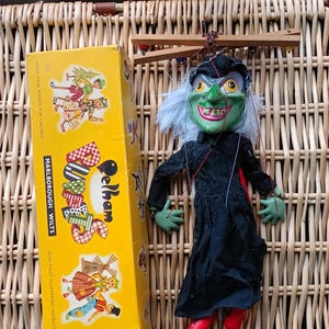 Puede incluir: Una marioneta de bruja Pelham vintage con una cara verde, un vestido negro y zapatos rojos, junto a su caja amarilla original. La caja presenta ilustraciones y el texto "Marlborough Wilts". La marioneta está suspendida de un control de madera.
