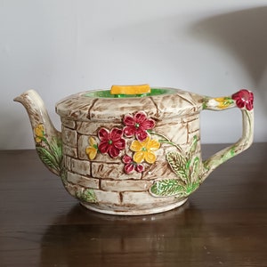 May include: A ceramic teapot with a brick-like design and floral accents. The teapot has a yellow lid and a brown handle. The flowers are red, yellow, and green.