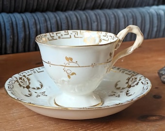 Puede incluir: Una taza de té y un platillo blancos con ribetes dorados y diseños florales. La taza tiene un asa y una base de pedestal. El platillo tiene un borde festoneado.