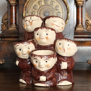 May include: A ceramic multi-cup holder featuring five sculpted figures with cream-colored faces and brown robes. Each figure has a distinct facial expression, including one wearing glasses and one with an open mouth, arranged in a tiered formation.