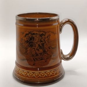 May include: A brown ceramic beer stein with a silver rim and handle. The stein features a black and white illustration of people riding horses. The bottom of the stein has a brown floral design.