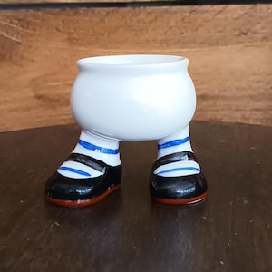 Puede incluir: Un huevero de cerámica blanca con piernas a rayas blancas y negras y zapatos negros con suelas rojas. El huevero está sobre una superficie marrón oscuro.