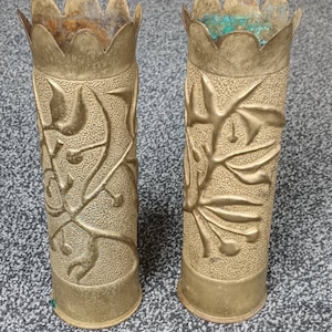 May include: Two brass vases with a decorative floral design. The vases are made from repurposed artillery shells and have a scalloped top.