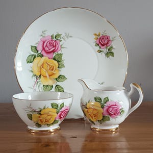 Pode incluir: Um conjunto de chá de porcelana vintage com um prato, um cremeira e um açucareiro. As peças brancas são adornadas com padrões florais de rosas amarelas e rosas e acabamentos dourados. O prato tem aproximadamente 23 cm de diâmetro.