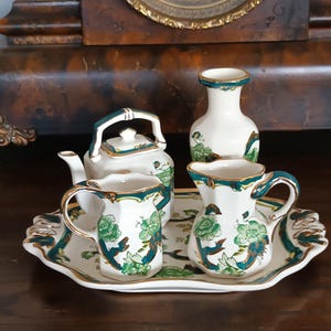 Puede incluir: Un juego de té vintage con una tetera, dos tazas, una jarra de crema y un jarrón, todo sobre una bandeja decorativa. El juego presenta una base blanca con diseños florales verdes y ribetes dorados. El fondo incluye un reloj de madera.