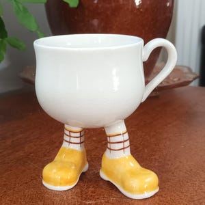 Puede incluir: Una taza de cerámica blanca con piernas y zapatos pintados de amarillo y marrón. La taza tiene un asa y tiene una forma tradicional.