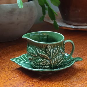 Op de afbeelding: Een groene keramische creamer en schotel set. De creamer heeft een tuit en een handvat, en beide stukken zijn versierd met een bladerenpatroon. De set staat op een houten oppervlak, met planten op de achtergrond.