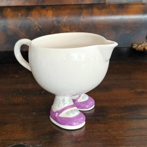 Puede incluir: Una cremera de cerámica blanca con asa y boquilla, apoyada sobre dos pies con forma de zapato morado. Los zapatos tienen suelas blancas y un diseño decorativo. La cremera está sobre una superficie de madera oscura.