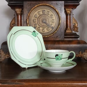 Puede incluir: Un juego de té vintage en verde claro y blanco. El juego incluye una taza, un platillo y un plato de forma única. El diseño presenta un patrón geométrico con detalles en negro. El juego se exhibe sobre una superficie de madera.