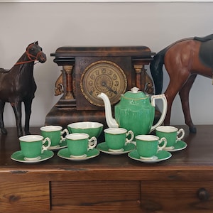Op de afbeelding: Een groen theeservies bestaande uit een theepot, een suikerpot en zes kopjes met bijpassende schoteltjes. Elk stuk heeft een textuur op het oppervlak met een gouden rand. De set staat voor een donkere houten klok en twee paardenbeeldjes.