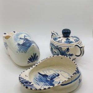Könnte beinhalten: Ein Set aus drei blau-weißen Keramikstücken im Delftware-Stil: ein Holzschuh, eine Teekanne und eine Schale. Die Stücke sind mit traditionellen holländischen Szenen verziert, darunter Windmühlen und Boote.